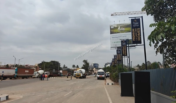 Wide view of the main access road leading towards the Prestige Evergreen development site, flanked by open landscapes and the project boundary hoardings under a cloudy sky.