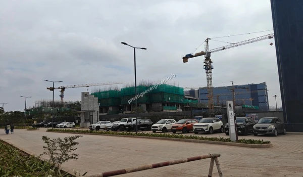 View of the ongoing structural development at the site, showing multi-story buildings covered in green safety mesh and tall tower cranes operating against the sky.